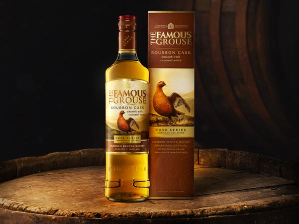 The Famous Grouse Bourbon Cask bottle and pack sitting on top of a barrel in a warehouse