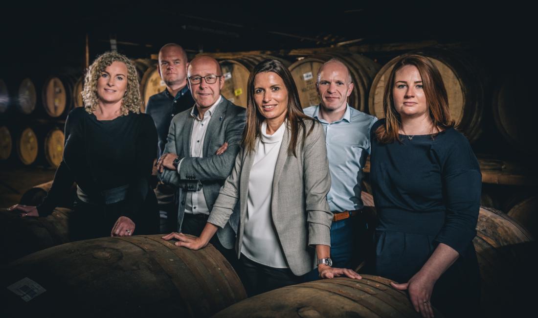 Group shot of three women and three men standing in front of casks in a warehouse