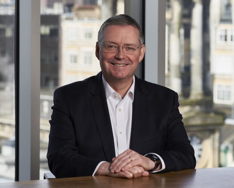 5x4 shot of CEO Scott McCroskie sitting at Boardroom table smiling with Glasgow skyline behind