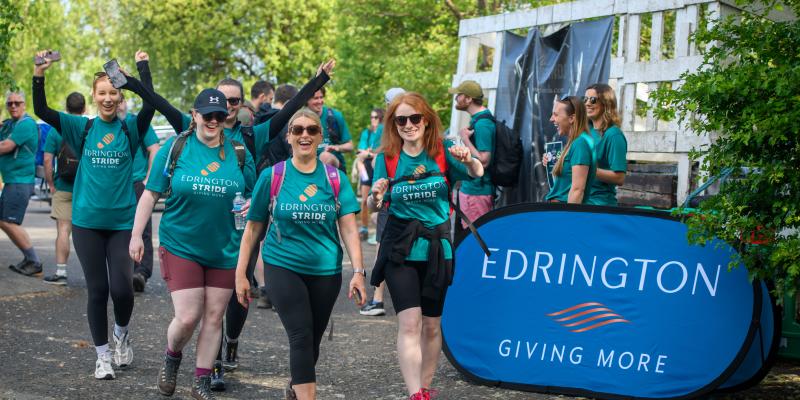 Four women walking along in teal Edrington Giving More t-shirts smiling and raising their hands as they walk past an Edrington Giving More sign