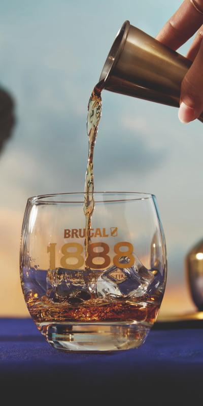 A hand pouring a measure of Brugal 1888 from a jigger into a Brugal 1888 branded glass